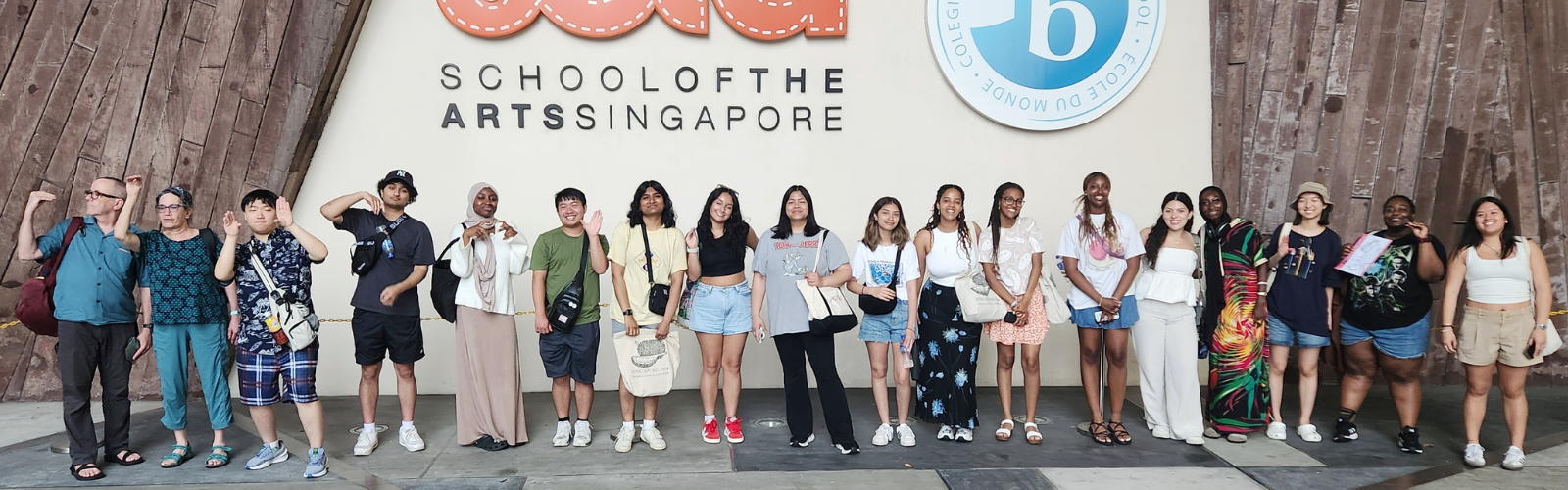 Sixteen students and two program leaders posed in a long line in front of a sign that says "SCHOOL OF THE ARTS SINGAPORE".
