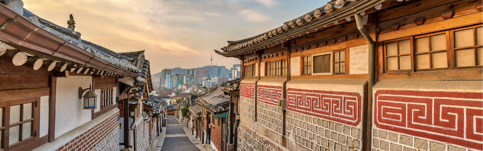 Narrow street lined with traditional Korean hanok houses featuring tiled roofs and ornate patterns. Cityscape and mountains in the background at sunset.