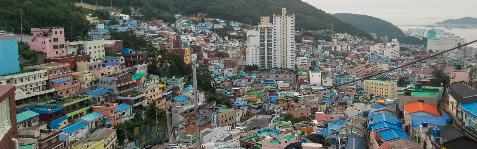Panoramic view of the hills of Seoul, South Korea, with colorful houses and high-rise buildings set against a backdrop of green hills, the ocean, and a cloudy sky.