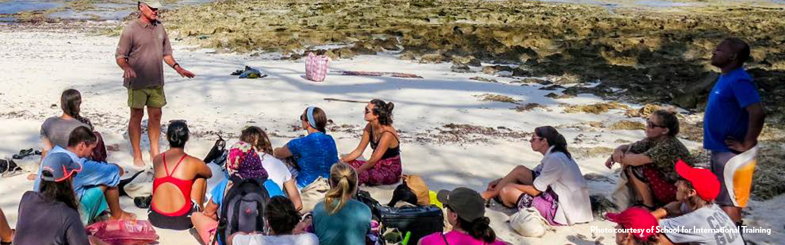 Program participants sitting on a beach, listening to a leader standing in front of them.