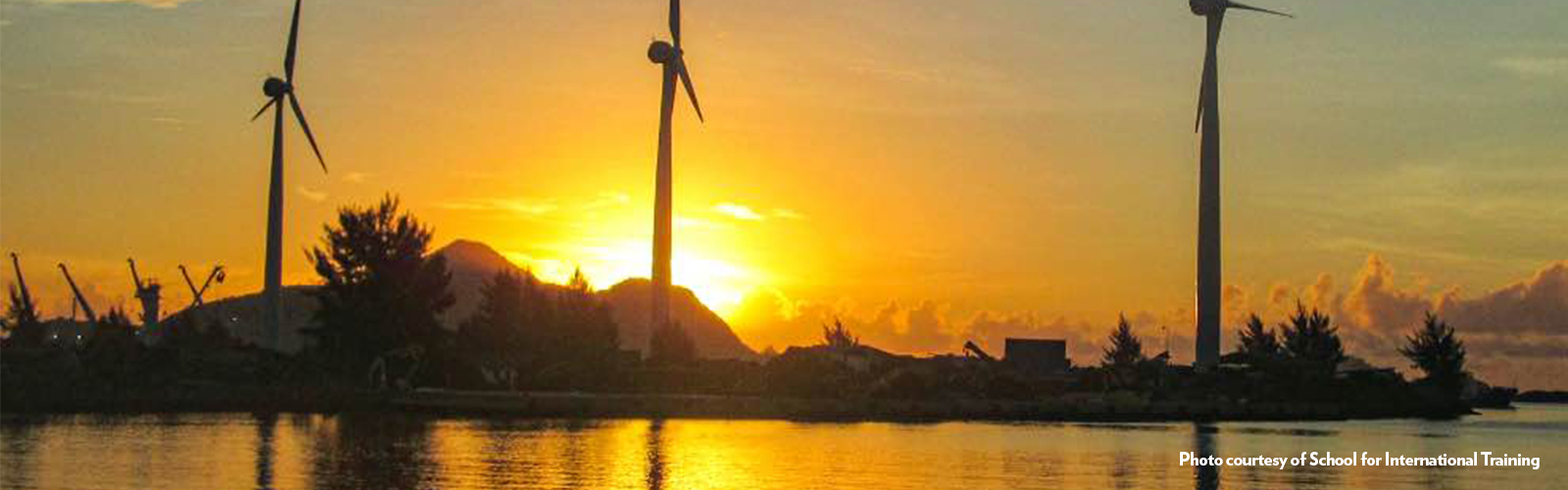 Three large wind turbines, near the water's edge, with the sun low on the horizon behind them.