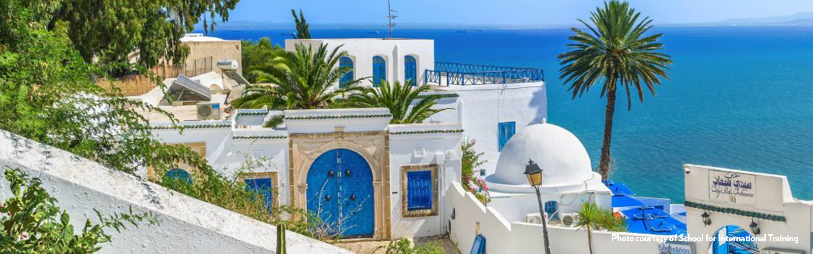 A white building with blue doors and windows, on a hill overlooking the sea.
