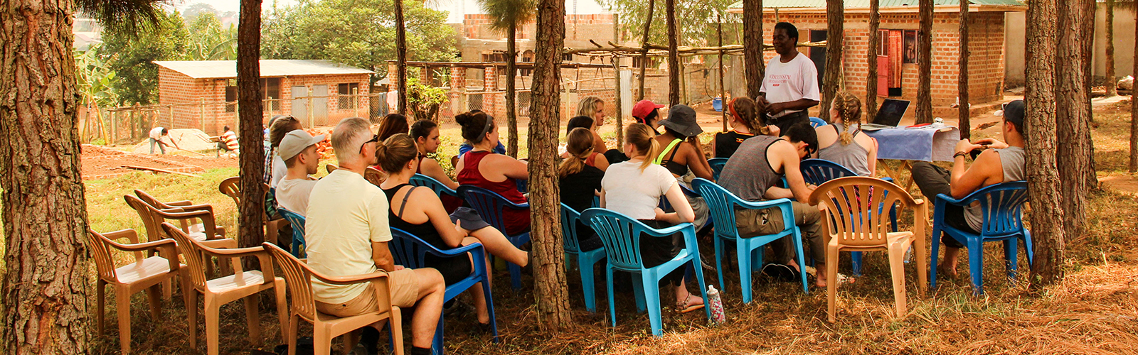 Program participants sitting outdoors on plastic chairs, listening to a speaker. Land is being farmed in the background behind them.