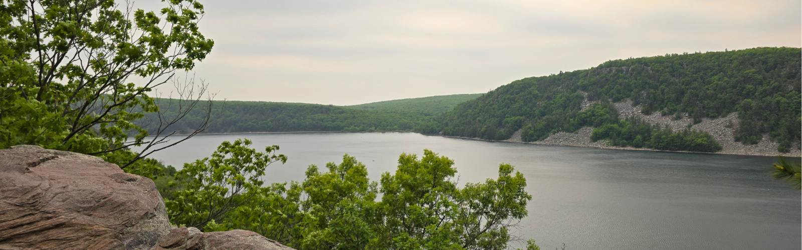 Devil's Lake bordered by lush green trees and rocky hills under an overcast sky.