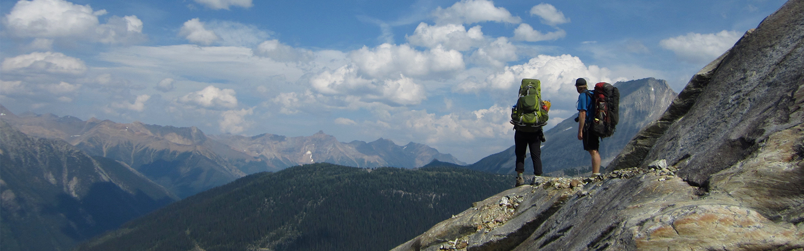 Two individuals wearing backpacking packs on a rocky ridge. Mountains line the horizon.