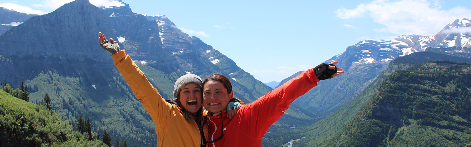 Two individuals posed at a point overlooking a wide valley flanked by snowy mountains.