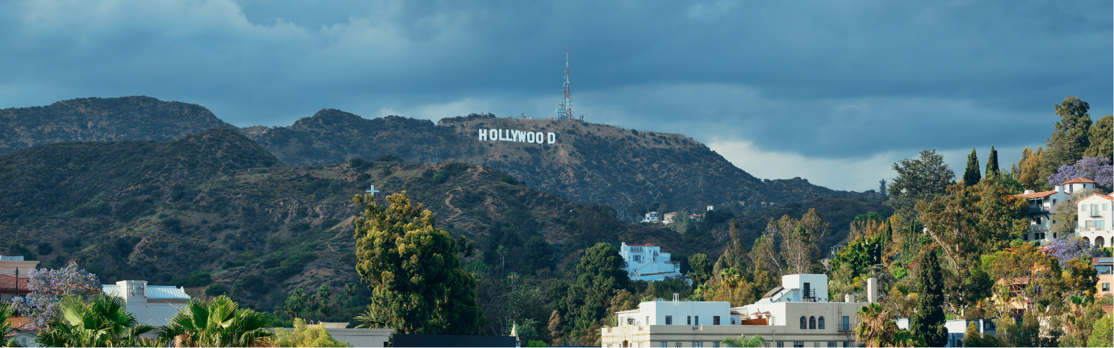 Cloudy sky over the iconic white Hollywood sign on a rugged hill surrounded by green vegetation and scattered houses.