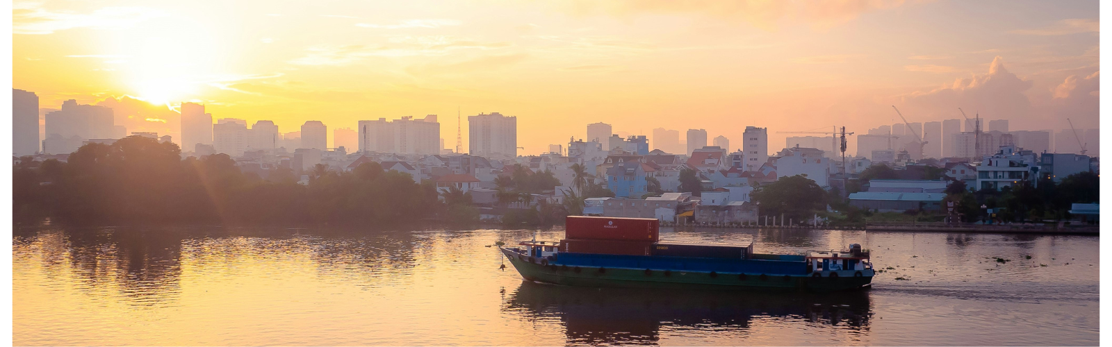 A medium-sized boat carrying several shipping containers down a waterway. Tall buildings line the horizon.